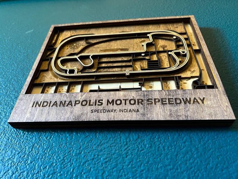 May include: A wooden wall art piece depicting the Indianapolis Motor Speedway race track. The track is made of layered wood and is surrounded by a dark brown frame. The text "Indianapolis Motor Speedway" and "Speedway, Indiana" is printed below the track.