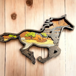 May include: A wooden horse silhouette with a layered design. The horse is running and the inside of the silhouette is filled with a scene of a forest with trees, a barn, and a river.
