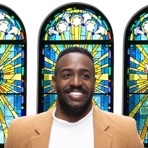 May include: A man with a brown jacket and a white shirt smiles in front of three stained glass windows. Each window features a white dove with yellow and blue accents.