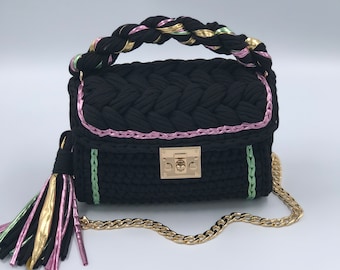May include: A black crocheted handbag with a braided handle and a gold chain strap. The bag has pink and green trim and a gold tassel.