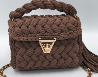 May include: A brown, hand-crocheted handbag with a braided handle and a gold-tone clasp. The bag features a gold chain strap and a tassel detail. The bag's texture is visible, showcasing the craftsmanship of the crochet work.
