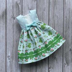 Könnte beinhalten: Ein hellblaues Puppenkleid mit einem Frosch- und Seerosenmuster. Das Kleid hat eine Schleife an der Taille und einen gewellten grünen Saum. Das Oberteil ist hellblau mit weißen Punkten.