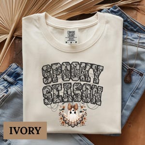 Op de afbeelding: Ivoorkleurige sweatshirt met de tekst "SPOOKY SEASON" in een zwart kanten ontwerp. Een cartoon spook met een strik en bloemaccenten staat onder de tekst. Het shirt wordt gepresenteerd op een bruin oppervlak met een blauwe jeans.