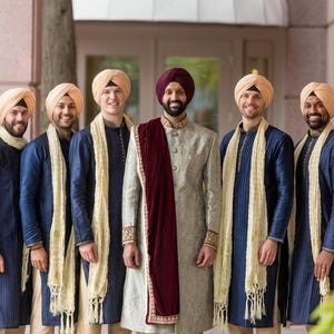 Puede incluir: Un grupo de nueve personas con vestimenta tradicional del sur de Asia, incluyendo sherwanis y turbantes. Ocho personas visten sherwanis azul marino con estolas color crema, mientras que la persona central viste un sherwani gris claro con un chal granate.