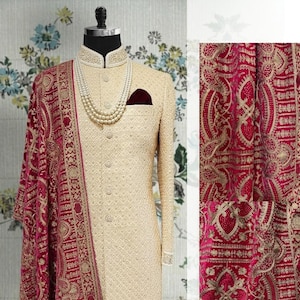 May include: A cream sherwani suit with detailed embroidery, a mandarin collar, and a pocket square. A maroon shawl with gold embroidery and tassels drapes over the suit. A pearl necklace adds elegance to the ensemble.