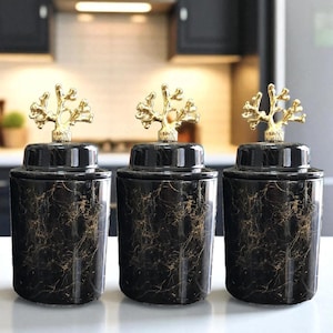 Puede incluir: Tres tarros decorativos negros con vetas doradas y tapas doradas en forma de coral. Los tarros son cilíndricos y se asientan sobre una superficie blanca. El fondo muestra un ambiente de cocina.