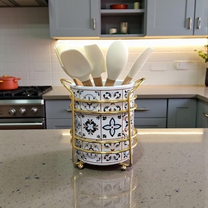 May include: A cylindrical ceramic utensil holder featuring a white base with a repeating blue, brown, and black geometric tile pattern. The holder is encased in a gold-toned wire frame with two side handles and contains several white silicone utensils.