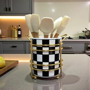 May include: A cylindrical ceramic utensil holder with a black and white chequered pattern, encased in a gold-toned wire frame with two side handles. The holder contains several off-white silicone kitchen utensils with light wooden handles.