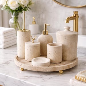 Op de afbeelding: Een beige badkameraccessoireset met een zeepdispenser, tandenborstelhouder en een potje met deksel. De set staat op een rond dienblad met gouden voetjes. Op de achtergrond een gouden kraan en spiegel.