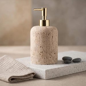 May include: A cylindrical soap dispenser made of beige, porous stone with a textured, pitted surface. The dispenser features a polished gold-tone metal pump mechanism at the top, designed for dispensing liquid soap or lotion with a simple press.