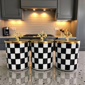 May include: Three kitchen canisters with a black and white chequered design. Each has a gold rim and a gold coral-shaped handle. The canisters are displayed on a speckled countertop in a kitchen setting.
