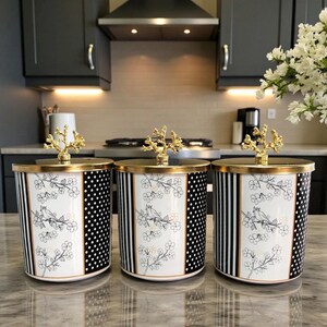 May include: Three decorative canisters with gold-colored lids and finials. The canisters have a black and white design with a bird and floral pattern. The canisters are displayed on a marble countertop.