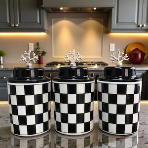Può includere: Tre barattoli abbinati a scacchi bianchi e neri con manici argentati a forma di albero sui coperchi. I barattoli cilindrici poggiano su un piano di lavoro riflettente. Sfondo della cucina.