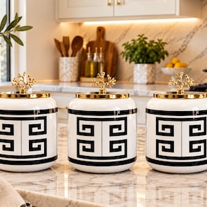 May include: A set of three white ceramic canisters featuring a bold black Greek key pattern around the centre. Each canister has a reflective gold lid topped with a decorative gold branch-like finial, creating a uniform look for kitchen storage.