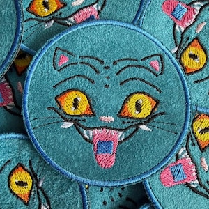 May include: A collection of teal, circular patches featuring a cartoon cat face. The cat has yellow eyes, a pink tongue, and pink and blue accents. The patches are embroidered with black thread.
