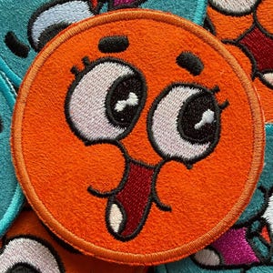 May include: Embroidered orange patch with a cartoon face featuring large white eyes, black pupils, and a red open mouth with a white tongue. The patch has a stitched border and is set against a background of similar patches.