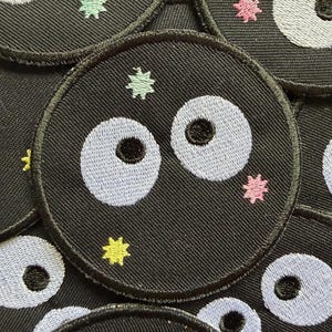 Könnte beinhalten: Eine Sammlung schwarzer, runder, gestickter Aufnäher. Jeder Aufnäher zeigt ein Cartoon-Gesicht mit hellblauen Augen und einer schwarzen Pupille. Kleine, farbenfrohe sternförmige Akzente in Rosa, Grün und Gelb umgeben die Gesichter. Die Aufnäher sind wahrscheinlich für Kleidung oder Accessoires.