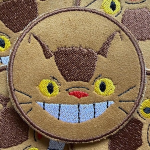 Peut inclure: Écussons brodés circulaires représentant un visage de chat souriant avec des yeux jaunes, un nez rouge et un large sourire avec des dents bleues. Les écussons sont bruns avec des accents bruns plus foncés.