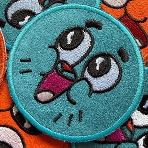 May include: Embroidered patch featuring a cartoon face with large, expressive eyes and a protruding tongue. The patch is teal with a teal border, and the face has black, white, and pink details. The design is whimsical and playful.