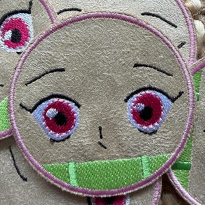 May include: Close-up of several embroidered patches featuring a cartoon character face. The patches have a beige background with pink and green accents. The eyes are large and red with white highlights, and the mouth is open.