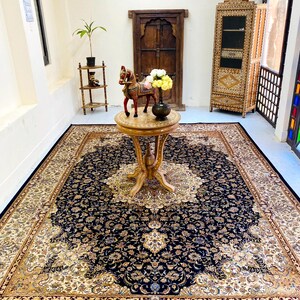 May include: A large, ornate area rug with a navy blue central field and a cream-colored border. The rug features an intricate floral pattern and is placed in a room with a wooden table and cabinet.