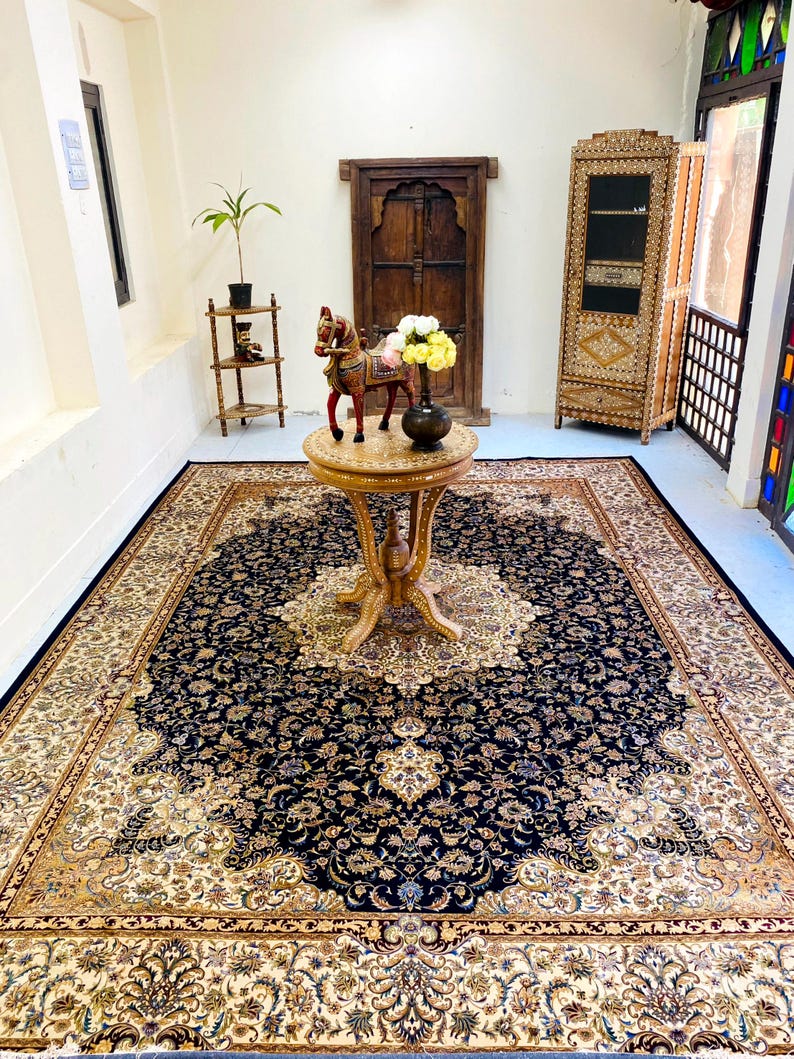 May include: A large, ornate area rug with a navy blue central field and a cream-colored border. The rug features an intricate floral pattern and is placed in a room with a wooden table and cabinet.