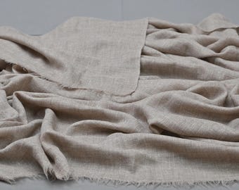 May include: A beige linen fabric, its textured weave creating soft folds and shadows. The edges are fringed. The natural colour and texture suggest a focus on comfort and simplicity, suitable for clothing or home textiles.