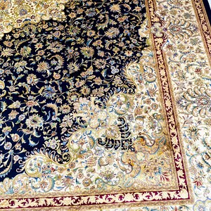 May include: An ornate, Persian-style rug with a navy blue field and a cream-colored border. The rug features intricate floral patterns in shades of gold, green, and burgundy. The design is symmetrical, with a central medallion and detailed corner elements.