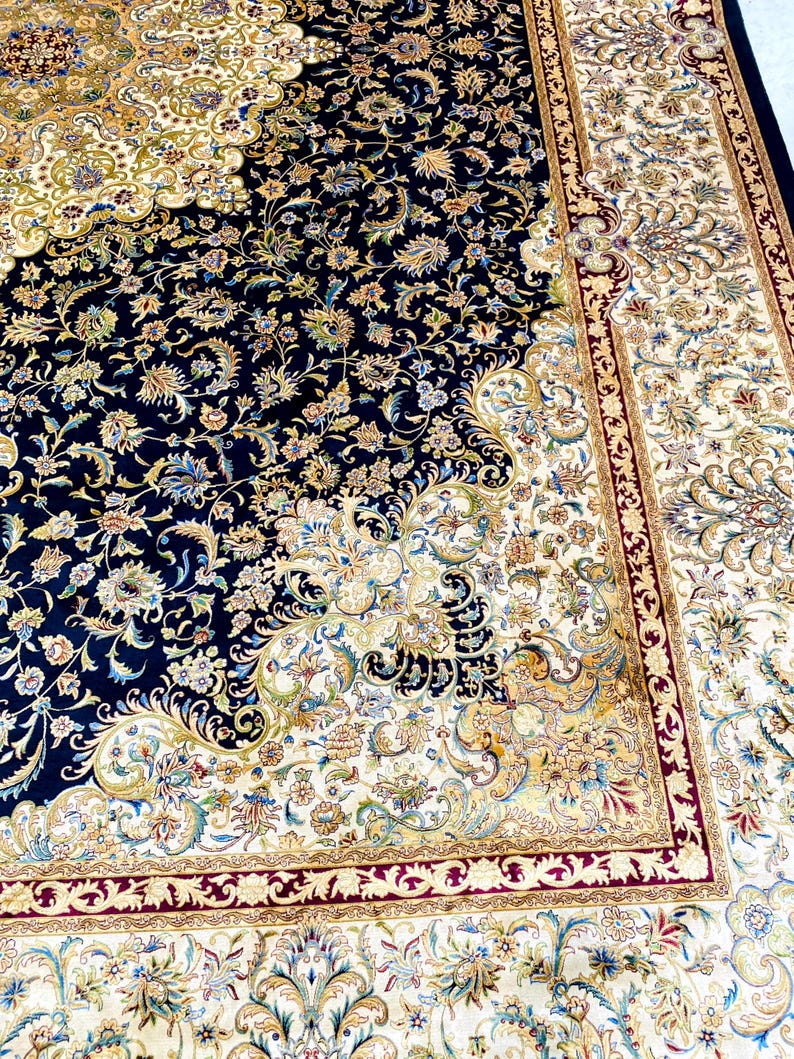 May include: An ornate, Persian-style rug with a navy blue field and a cream-colored border. The rug features intricate floral patterns in shades of gold, green, and burgundy. The design is symmetrical, with a central medallion and detailed corner elements.