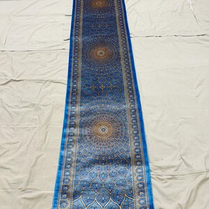 May include: A long, rectangular blue runner rug with an intricate, symmetrical pattern. The rug features a central design with circular medallions and a border with repeating motifs. The color palette includes shades of blue, gold, and cream.