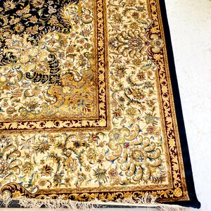 May include: A detailed shot of a Persian rug with a navy blue field and a cream-colored border. The rug features intricate floral patterns in gold, cream, and burgundy. The rug has a black border and a fringed edge.