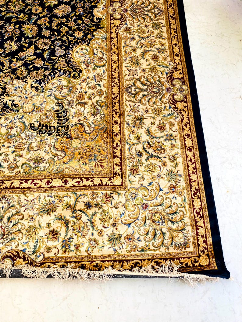 May include: A detailed shot of a Persian rug with a navy blue field and a cream-colored border. The rug features intricate floral patterns in gold, cream, and burgundy. The rug has a black border and a fringed edge.