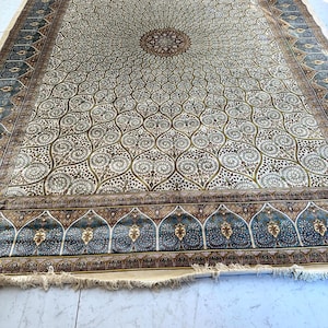 May include: An ornate, rectangular rug with a central medallion design. The rug features a cream-colored field with a repeating pattern of floral motifs in shades of brown and green. A border of blue and brown frames the rug, with a fringe along the edge.