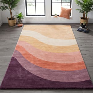 May include: A rectangular area rug with a wavy, layered design in shades of purple, pink, orange, and beige. The rug is made of a soft, plush material and is suitable for a living room or bedroom. The rug's dimensions are not visible in the image.