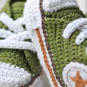 以下が含まれることがあります： 白のひもで結ばれた緑色のベビーシューズ。茶色とオレンジの縁取りと、つま先に白い星が描かれています。