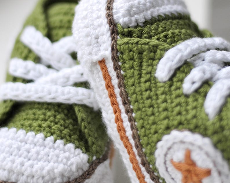 以下が含まれることがあります： 白のひもで結ばれた緑色のベビーシューズ。茶色とオレンジの縁取りと、つま先に白い星が描かれています。