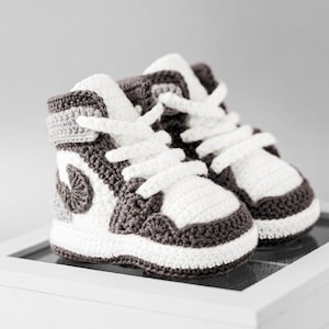 K&ouml;nnte beinhalten: Ein Paar handgefertigte Baby-Sneaker aus H&auml;kelarbeit im High-Top-Stil. Die Schuhe sind wei&szlig;, grau und dunkelbraun mit wei&szlig;en Schn&uuml;rsenkeln. Die Sneaker werden auf einer wei&szlig;en Oberfl&auml;che pr&auml;sentiert.