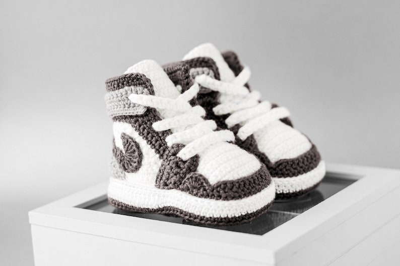 K&ouml;nnte beinhalten: Ein Paar handgefertigte Baby-Sneaker aus H&auml;kelarbeit im High-Top-Stil. Die Schuhe sind wei&szlig;, grau und dunkelbraun mit wei&szlig;en Schn&uuml;rsenkeln. Die Sneaker werden auf einer wei&szlig;en Oberfl&auml;che pr&auml;sentiert.