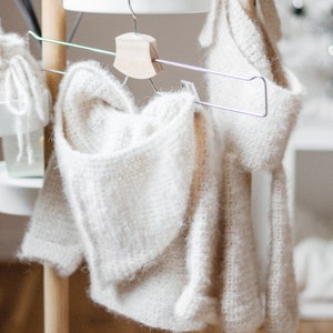 Pode incluir: Duas pe&ccedil;as de roupa de malha branca penduradas em um rack de madeira com uma prateleira branca. As pe&ccedil;as s&atilde;o um su&eacute;ter com capuz e uma echarpe.