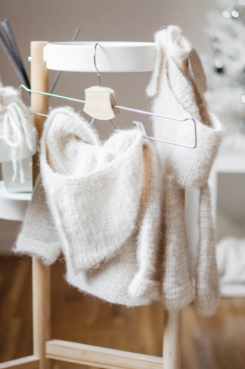 Pode incluir: Duas pe&ccedil;as de roupa de malha branca penduradas em um rack de madeira com uma prateleira branca. As pe&ccedil;as s&atilde;o um su&eacute;ter com capuz e uma echarpe.