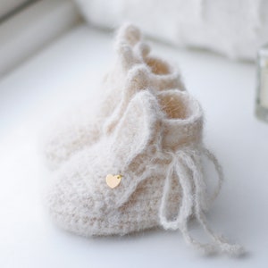 Pode incluir: Um par de botinhas de beb&ecirc; de tric&ocirc; cor creme com textura felpuda e um pequeno charme de cora&ccedil;&atilde;o dourado. As botinhas t&ecirc;m um la&ccedil;o na parte superior.
