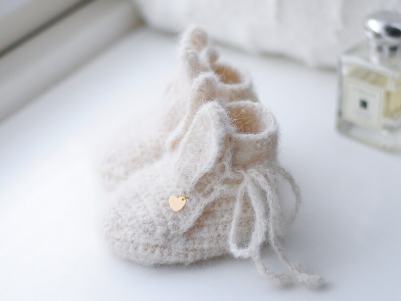 Pode incluir: Um par de botinhas de beb&ecirc; de tric&ocirc; cor creme com textura felpuda e um pequeno charme de cora&ccedil;&atilde;o dourado. As botinhas t&ecirc;m um la&ccedil;o na parte superior.