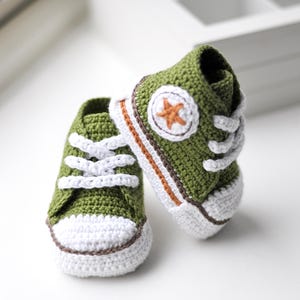 以下が含まれることがあります： 緑色の毛糸で編まれたベビーシューズのペア。白い紐と、側面に茶色の星が付いています。