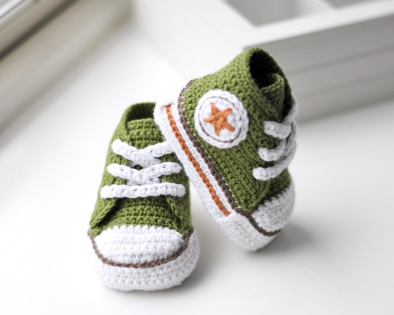 以下が含まれることがあります： 緑色の毛糸で編まれたベビーシューズのペア。白い紐と、側面に茶色の星が付いています。