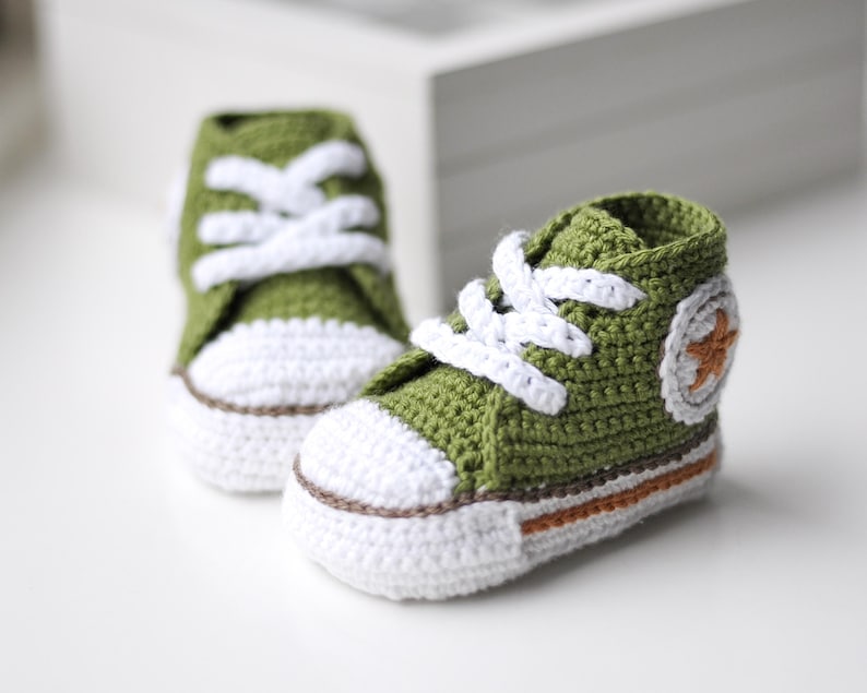以下が含まれることがあります： 白のレースと側面に白い星が付いた緑色のクロシェ編みのベビーシューズのペアです。