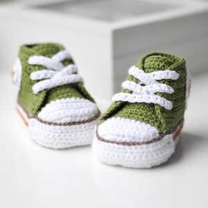 以下が含まれることがあります： 緑色の編みぐるみベビーシューズのペア。白い靴ひも、白いソール。クラシックなスニーカーデザインです。