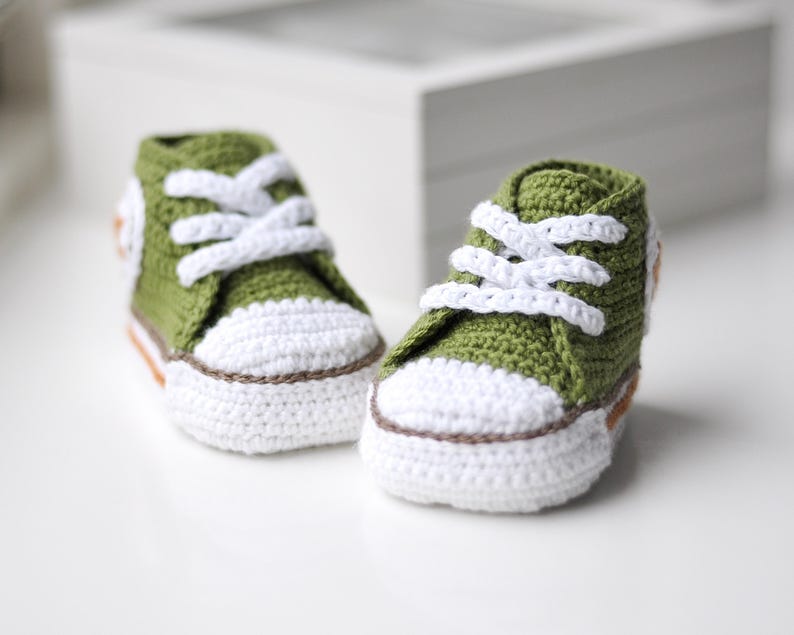 以下が含まれることがあります： 緑色の編みぐるみベビーシューズのペア。白い靴ひも、白いソール。クラシックなスニーカーデザインです。