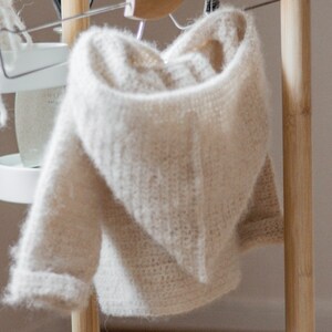 Pode incluir: Um su&eacute;ter de croch&ecirc; com capuz de cor creme pendurado em um cabide de madeira.