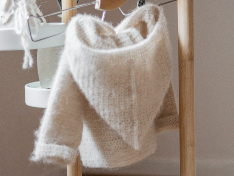 Pode incluir: Um su&eacute;ter de croch&ecirc; com capuz de cor creme pendurado em um cabide de madeira.