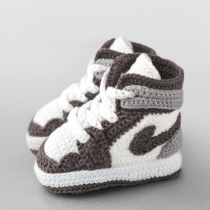 K&ouml;nnte beinhalten: Ein Paar handgefertigte Baby-H&auml;kel-Sneaker im High-Top-Stil. Die Schuhe haben eine wei&szlig;e Basis mit braunen und grauen Akzenten, einschlie&szlig;lich des ikonischen Swoosh-Designs. Wei&szlig;e Schn&uuml;rsenkel vervollst&auml;ndigen den Look. Ein einzigartiges Geschenk.